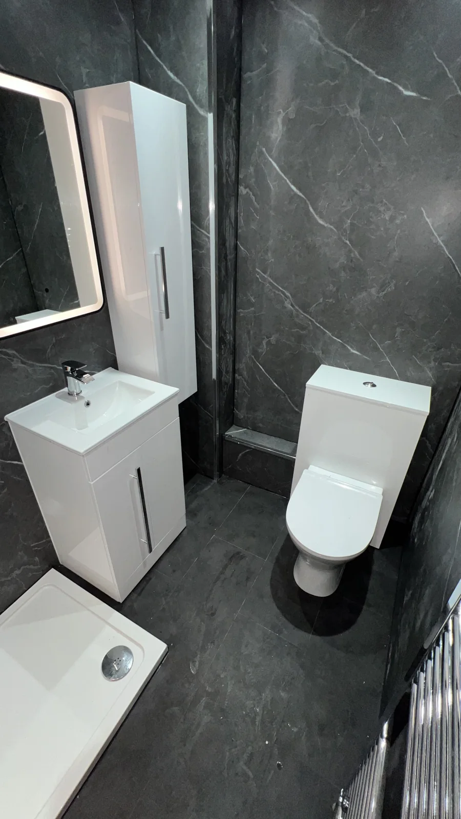 White basin vanity, tall storage cupboard and toilet against dark marble-effect walls