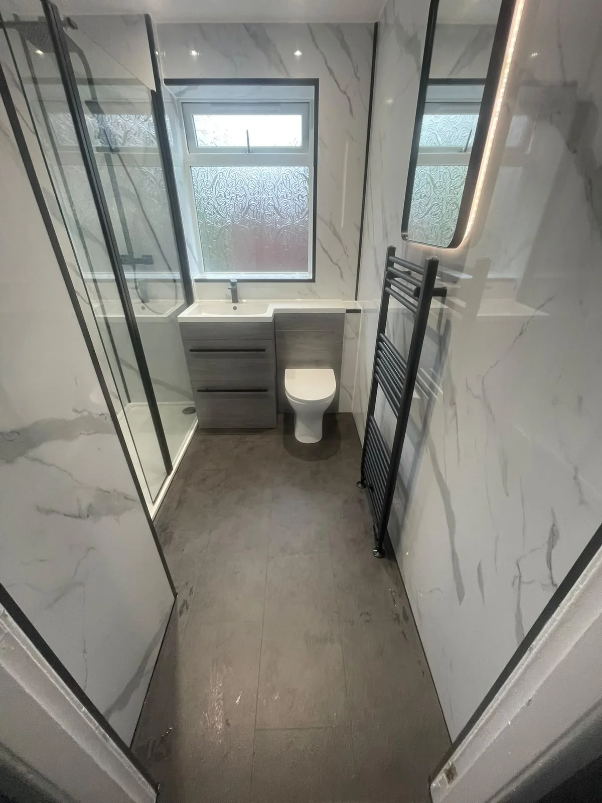 Narrow shower room with grey wood-effect vanity storage and a black towel radiator