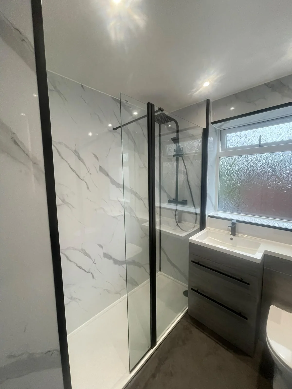 Glass shower enclosure beside grey vanity drawers and a white toilet